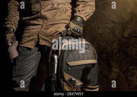 Uomo in giacca da tempesta e guanti militari tattici che tengono uno zaino con attrezzatura da viaggio su fondo parete in pietra. Foto Stock