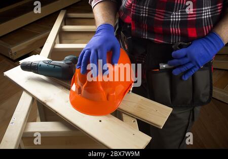 Operaio in guanti blu, camicia nera e rossa che tiene un portautensili professionale e casco da costruzione posato su una mensola di legno con trapano. Foto Stock