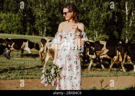 Ritratto esterno atmosfera stile di vita foto di giovane bella donna dai capelli scuri in un abito rosa si erge sullo sfondo di un gregge di mucche. Ragazza Foto Stock