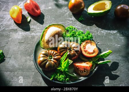 Avocado, pomodori e altre verdure crude su piatto nero, vista dall'alto. Concetto di cibo sano Foto Stock