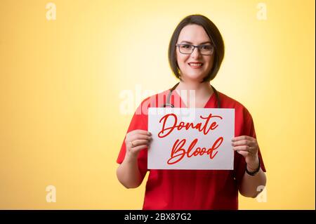 Ritratto di bella donna dottore con stetoscopio con scrub rossi, tenendo una carta bianca e sorridendo posando su uno sfondo isolato giallo. Foto Stock