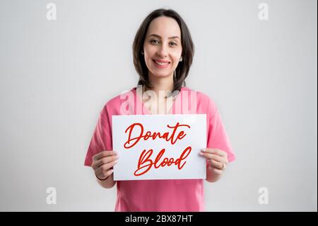 Smilieng donna medico infermiere o medico vestito con abiti rosa ospedale, con capelli marroni, tenendo una carta con messaggio donare sangue pos Foto Stock