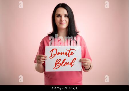 Ritratto di bella donna dottore con stetoscopio con scrub rosa, tenendo una carta bianca e sorridendo posando su un rosa isolato retrogusto. Foto Stock
