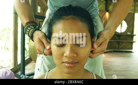 Primo piano ritratto di giovane bella e rilassata donna indonesiana asiatica che riceve tradizionale massaggio facciale tailandese con le mani maschili che lavorano le orecchie Foto Stock