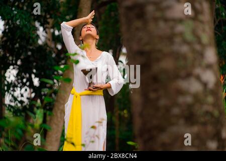 Connessione mente e corpo - bella e felice guaritrice donna asiatica tenendo incenso tazza facendo rituale tradizionale danza guarigione a verde foresta tropicale Foto Stock