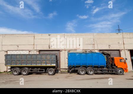 Camion grande con cabina arancione, un corpo blu e un rimorchio grigio contro un deposito di cemento con un cancello di ferro chiuso e il cielo in una giornata di sole attesa f Foto Stock