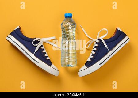 Sneaker o scarpe da ginnastica classiche blu con lacci per scarpe bianchi e bottiglia di acqua potabile su sfondo giallo testurizzato. Scarpe per il fitness e lo sport. Foto Stock