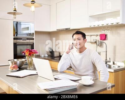 giovane uomo d'affari asiatico seduto cucina contatore che lavora a casa parlando sul telefono cellulare Foto Stock