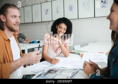 Ritratto di bella ragazza afroamericana sorridente con capelli ricci scuri seduti con amici in ufficio. Gruppo di studenti che parlano mentre studiano il Tog Foto Stock