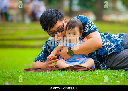 Felice e giocoso giapponese asiatico uomo come padre amorevole godendo con dolce e bella bambina figlia seduta insieme giocando sull'erba alla città p Foto Stock