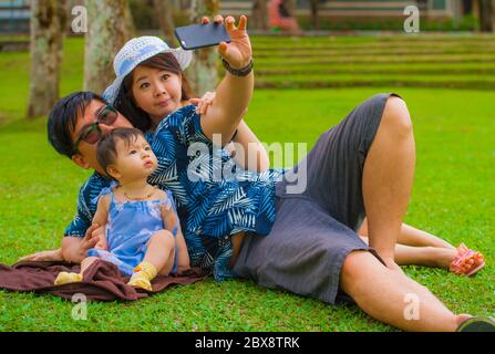 Giovane felice e amorevole famiglia asiatica coreana con genitori e dolce figlia bambino al parco della città insieme con il padre che prende foto selfie con il telefono cellulare Foto Stock