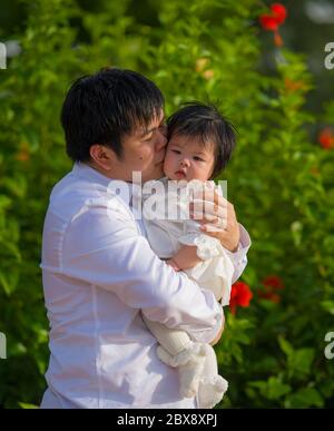 giovane uomo felice e orgoglioso come padre di bambina dolce che tiene la figlia di fronte al giardino dei fiori al resort vacanze godendo insieme Foto Stock