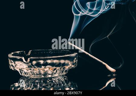 Un sigaro che brucia in un posacenere di vetro con fumo visibile su sfondo nero. Studio girato . Foto tasto basso . Foto Stock