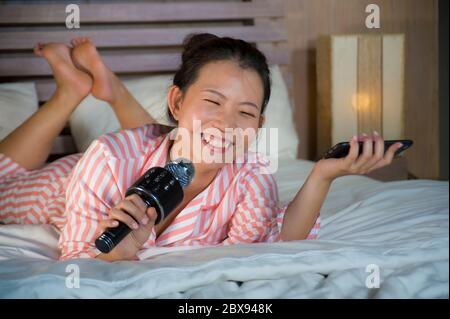 Giovane felice e bella Asian ragazza teenager cinese che canta karaoke canzone eccitato a casa camera da letto tenendo telefono cellulare giocare a letto eccitato e che Foto Stock