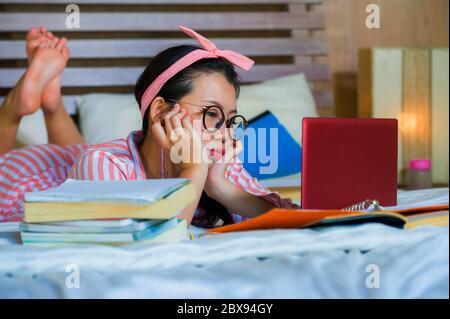 Giovane cute e felice nerdy asiatico studente coreano adolescente donna in nerd capelli nastro studiare a casa camera da letto seduta sul letto con computer portatile e te Foto Stock