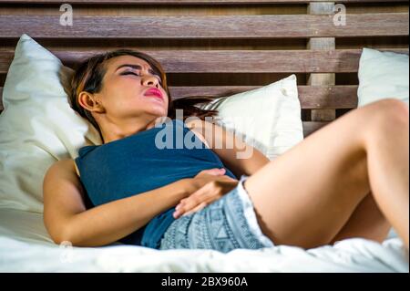 Giovane attraente Asian Latina mista donna sdraiata sul letto che soffre mal di crampi allo stomaco e dolore di periodo sensazione di malessere e malessere nel ciclo e doloroso menstr Foto Stock