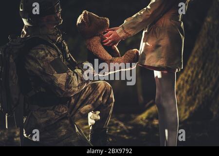Tema esercito e patriottismo. Soldato che dà il giocattolo dell'orso del Teddy alla ragazza caucasica nella foresta scura del Foggy Foto Stock