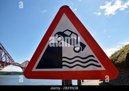 Primo piano del cartello con il triangolo di avvertimento per l'auto che cade in acqua con il Forth Rail Bridge sfocato sullo sfondo, giorno di sole con cielo blu e nuvola di luce Foto Stock