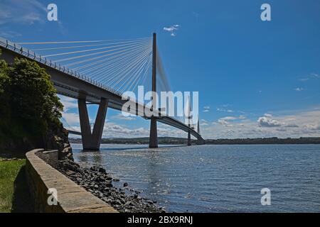 Queensferry Crossing in Scozia, Regno Unito in una giornata di sole con cielo blu e nuvole. Parete di mare, rocce, erba e alberi in vista. Foto Stock