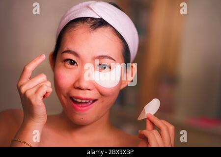 Giovane bella e felice asiatica cinese donna sorridente a bagno specchio applicando idratante occhi patch sotto gli occhi per riparare cerchi scuri in beaut Foto Stock