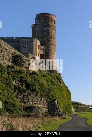 La torre del Castello di Bambburgh e il muro sopra il parco aperto e il sentiero a Bambburgh in Northumberland, Inghilterra, Regno Unito sotto il cielo azzurro chiaro e illuminato dal sole. Foto Stock