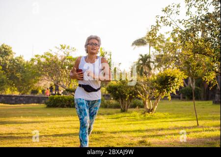 medio anni 40 o 50 donna felice e attraente con capelli grigi allenamento al parco cittadino con alberi verdi all'alba facendo corsa e jogging allenamento i Foto Stock