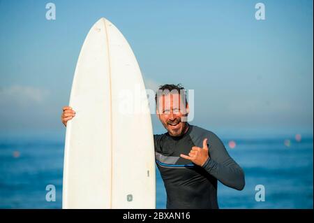 Stile di vita ritratto sulla spiaggia di bello e attraente uomo surfista in neoprene swimsuit che tiene surf tavola in posa fresco dopo il surf godendo di acqua estiva Foto Stock