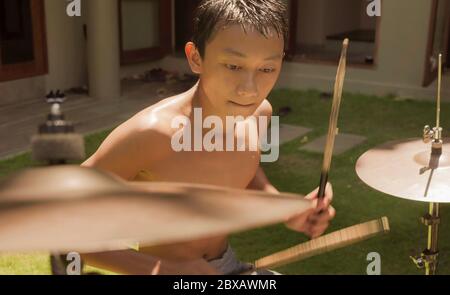 Teenager asiatico americano che gioca dums. Ritratto estivo di bel ragazzo che pratica su drum kit a casa giardino prova rock canzone provando il suo ho Foto Stock