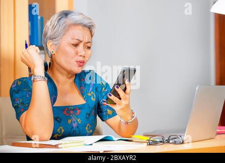 ritratto di stile di vita dell'ufficio di attraente donna asiatica preoccupata e sollecitata di mezza età che usa il telefono mobile alla scrivania del computer portatile che si sente sopraffatta Foto Stock