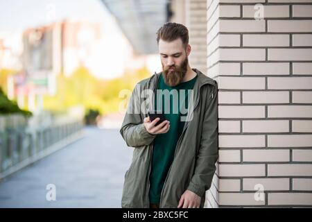 Bell'uomo brutale bearded sta usando il telefono sulla strada soleggiata Foto Stock