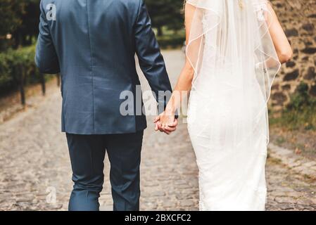 Vista posteriore della coppia sposata che cammina su una strada lastricata di pietra. Sposo e sposa che tengono le mani. Concetto di giorno di nozze. Foto Stock