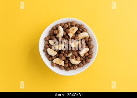 Ciotola bianca piena di palle di cereali al cioccolato e fette di banana su sfondo giallo, vista dall'alto verso il basso. Moderna e vivace immagine della colazione. Foto Stock
