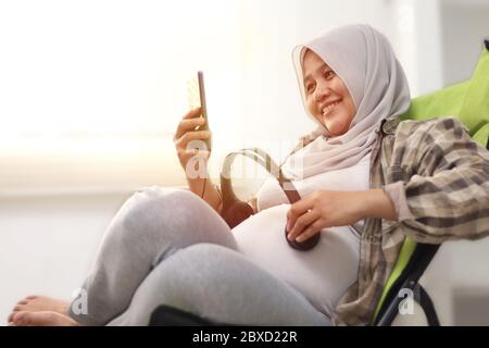 Bella musulmana asiatica incinta mette le cuffie sul suo pancino e apre la recitazione di Quran o musica classica per il suo bambino ascolto. Felice e rilassante Foto Stock