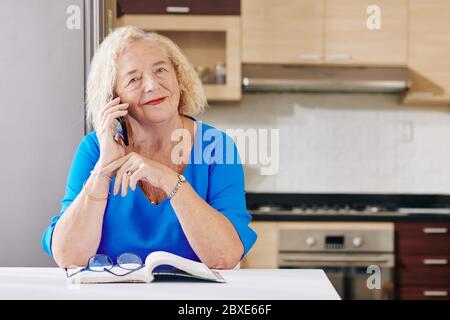 Donna positiva anziana seduta a tavola in cucina e chiamando i suoi amici o parenti Foto Stock