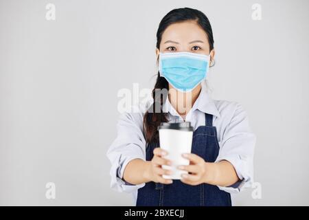 Bella barista femminile in maschera medica dando tazza di caffè take away ha fatto per il cliente Foto Stock