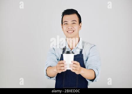 Giovane barista asiatico allegro che dà una tazza di caffè da asporto, isolato su grigio Foto Stock