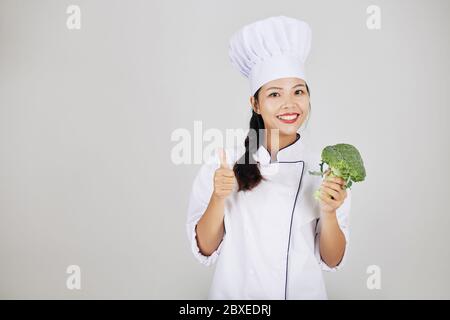 Ritratto di giovane cuoco felice che tiene freschi broccoli e mostra pollici-su Foto Stock