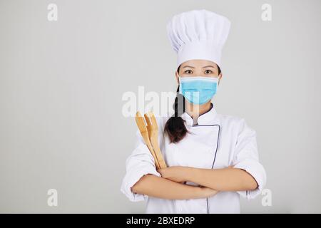 Giovane chef di fiducia in maschera medica posa con utensili in legno Foto Stock