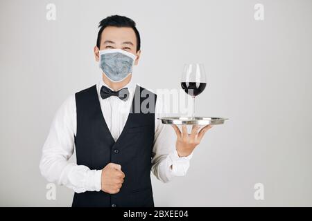 Ritratto di cameriere felice ristorante in maschera medica che serve un bicchiere di vino rosso Foto Stock