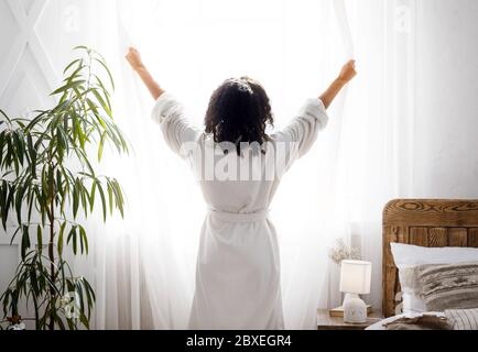 Mattina perfetta. Donna irriconoscibile in accappatoio bianco che apre le tende della finestra a casa Foto Stock