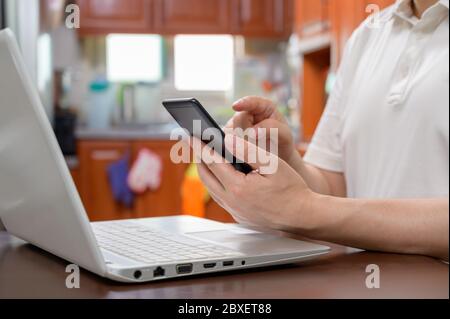 Un uomo di mezza età vestito casualmente a casa e lavora da casa utilizzando uno smartphone e un computer. Connessione rete Internet, connessione telelavoro Foto Stock