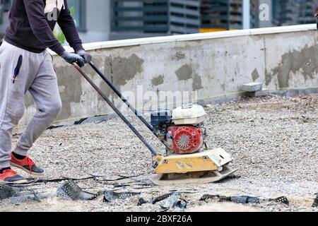 Un lavoratore di strada usa una stufa a benzina per compattare pietra frantumata al luogo di riparazione stradale e successiva posa di lastre di pavimentazione. Foto Stock