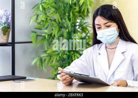 Ritratto di donna asiatica medico indossare maschera protettiva viso sedersi nella sua stanza di ufficio in clinica ospedale e leggere sul file di pazienza prima di controllare patie Foto Stock