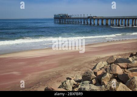 Molo sull'Oceano Atlantico a Swakompund, Namibia. Bella sabbia rosa, onde e pietre in una giornata soleggiata e luminosa. Foto Stock