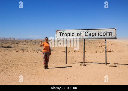 Giovane viaggiatore caucasico con capelli rossi zaino in spalla sullo sfondo di un cartello stradale Tropic di Capricorno nel deserto del Namib, Namibia, Africa. Foto Stock