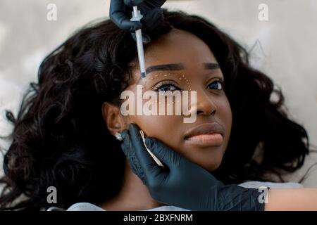 Primo piano di donne cosmetologist medico mani in guanti neri facendo iniezioni biorevitalizzazione in bella donna africana faccia pelle intorno agli occhi Foto Stock