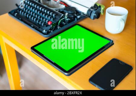 accanto a una vecchia macchina da scrivere si trova una tavoletta con uno schermo verde sul tavolo, un telefono cellulare e un caffè Foto Stock