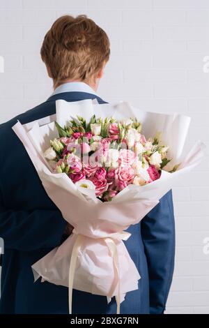 giovane biondo in tuta blu tiene in mano un grande bouquet di rose bianco-rosa ed eustoma sullo sfondo bianco della parete Foto Stock