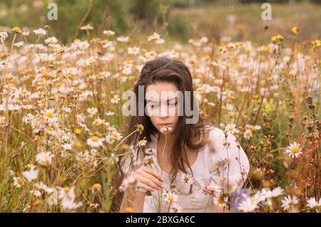 Giovane bella ragazza in abito bianco che odora fiori in camomilla campo in estate Foto Stock