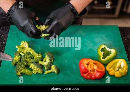Primo piano di chef irriconoscibile in guanti neri che preparano verdure fresche sul tagliere Foto Stock